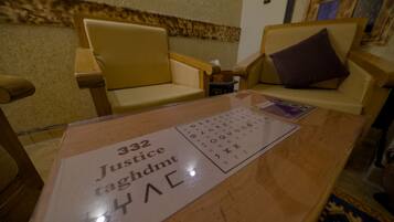 Ein Zimmer mit einem Holztisch, zwei beigen Sesseln und einem kleinen Tisch mit einem Schild, auf dem '332 Justice taghdmt' steht.