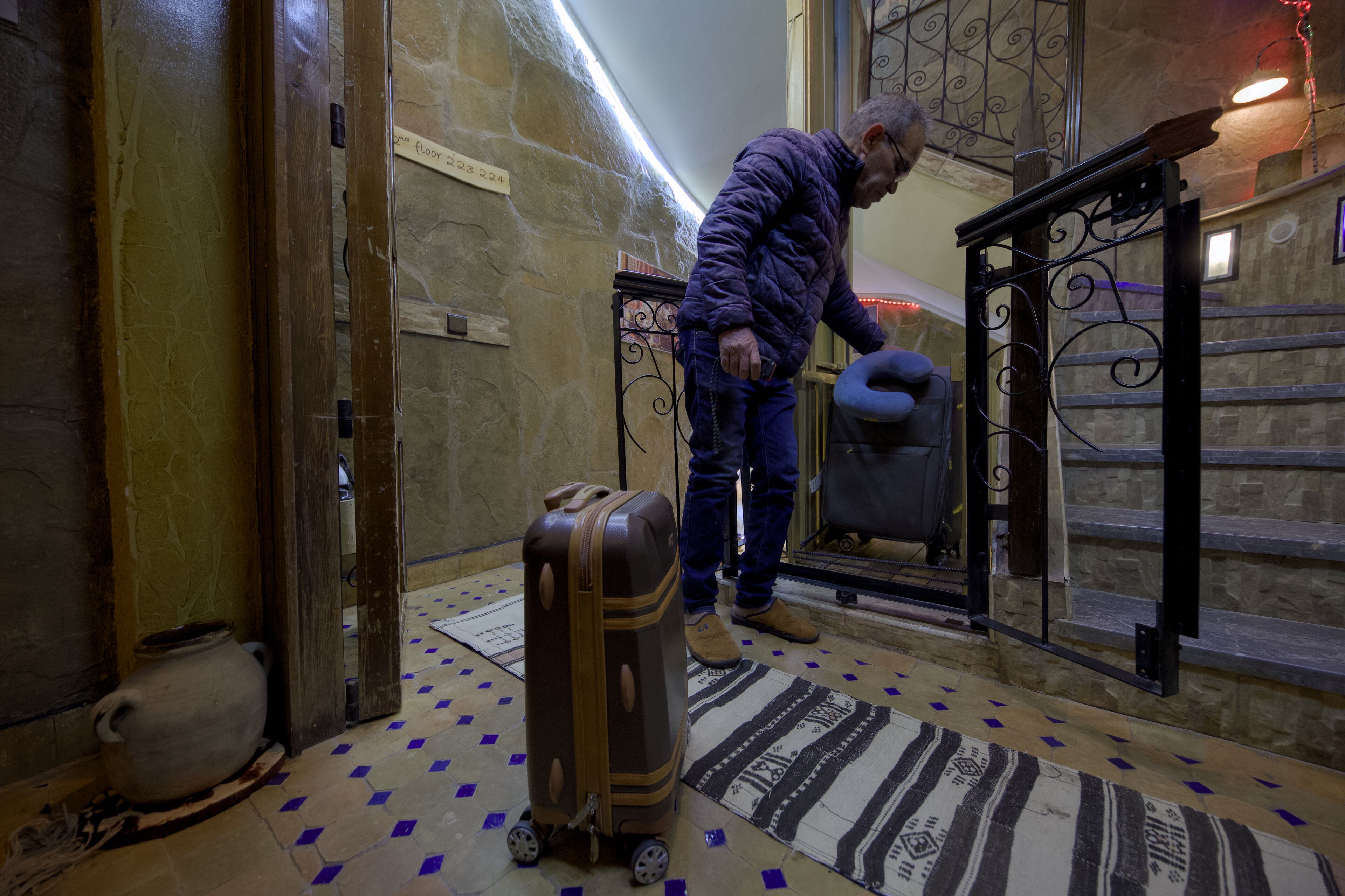 Un hombre con una chaqueta púrpura y jeans está de pie junto a una maleta en una habitación con piso estampado y pared de piedra.