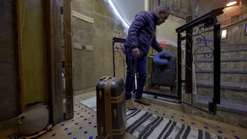 Un hombre con una chaqueta púrpura y jeans está de pie junto a una maleta en una habitación con piso estampado y pared de piedra.