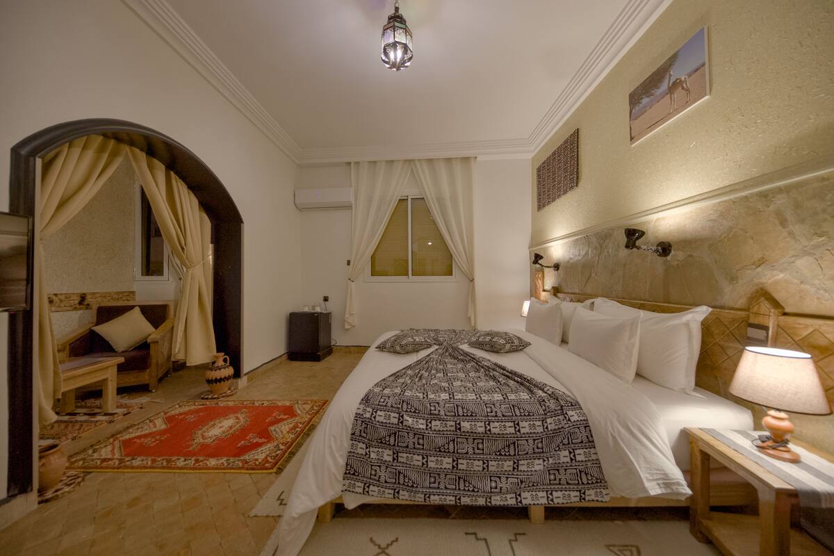 A bedroom with a bed, a wooden bench, a red rug, a lamp, and a wall-mounted picture.