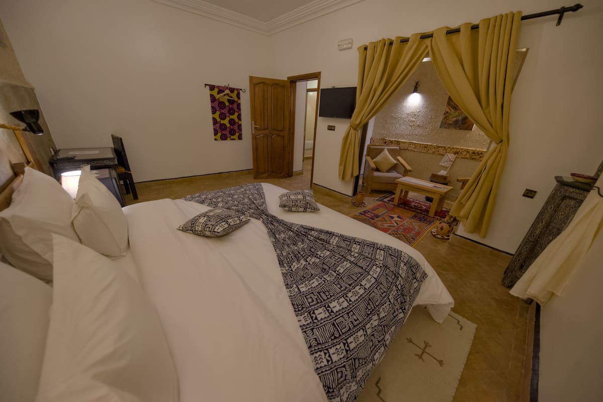 A bedroom with a large bed, a desk, a chair, a TV, and a bathroom visible through a door.