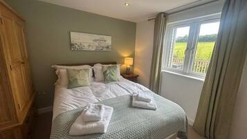 Una cama bien tendida con una colcha verde estampada y dos toallas dobladas, una lámpara de noche y una ventana con vistas a un campo verde.