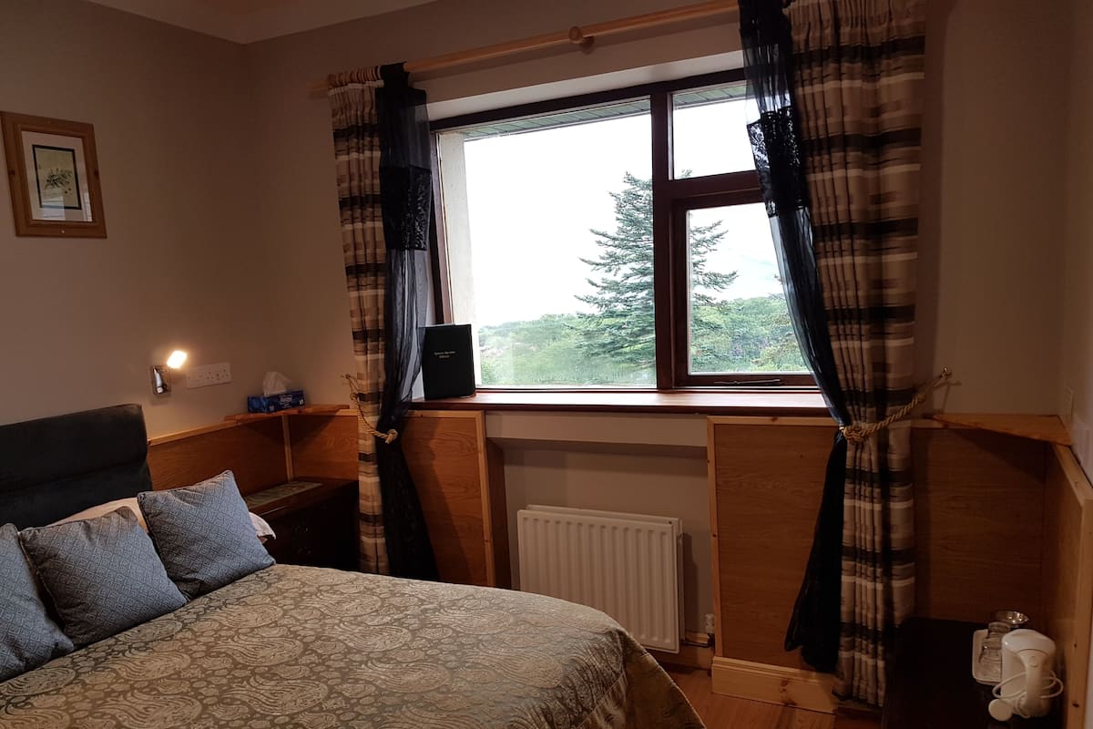 A bedroom with a bed, a window with curtains, a radiator, and a small table with a lamp.