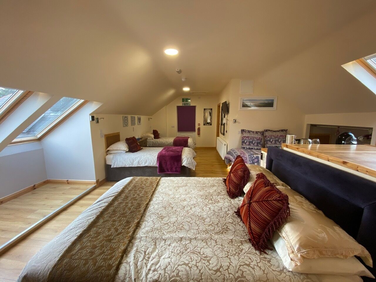 A spacious room with a large bed, a sofa, and a dining table. There are skylights on the ceiling, and a staircase is visible in the background.