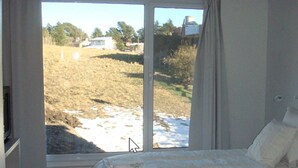Une chambre avec une grande fenêtre, un lit recouvert de draps blancs et une vue sur un champ verdoyant avec un bâtiment visible au loin.