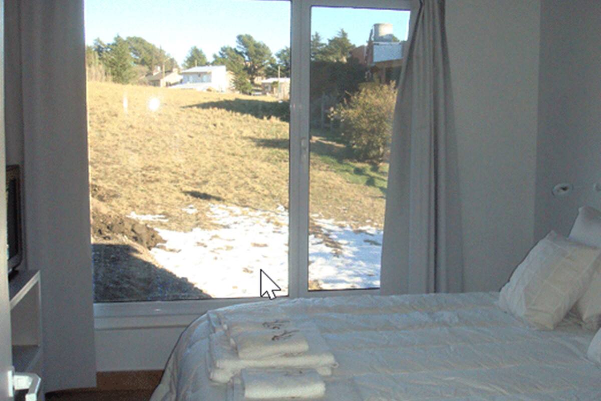 A bedroom with a large window, a bed with white linens, and a view of a grassy field with a building in the distance.
