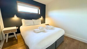 A neatly made bed with white linens and towels, a bedside table with a lamp, and a window above the bed.