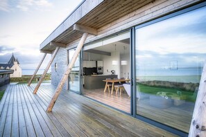 A modern house with a wooden deck, a dining area with a table and chairs, and a kitchen visible through glass doors.