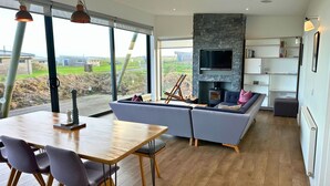 A modern living room with a stone fireplace, a sofa, a dining table with chairs, and a large window offering a view of the outdoors.
