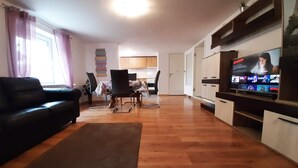 A living room with a black sofa, a dining table with chairs, a TV on a stand, and a kitchen in the background.