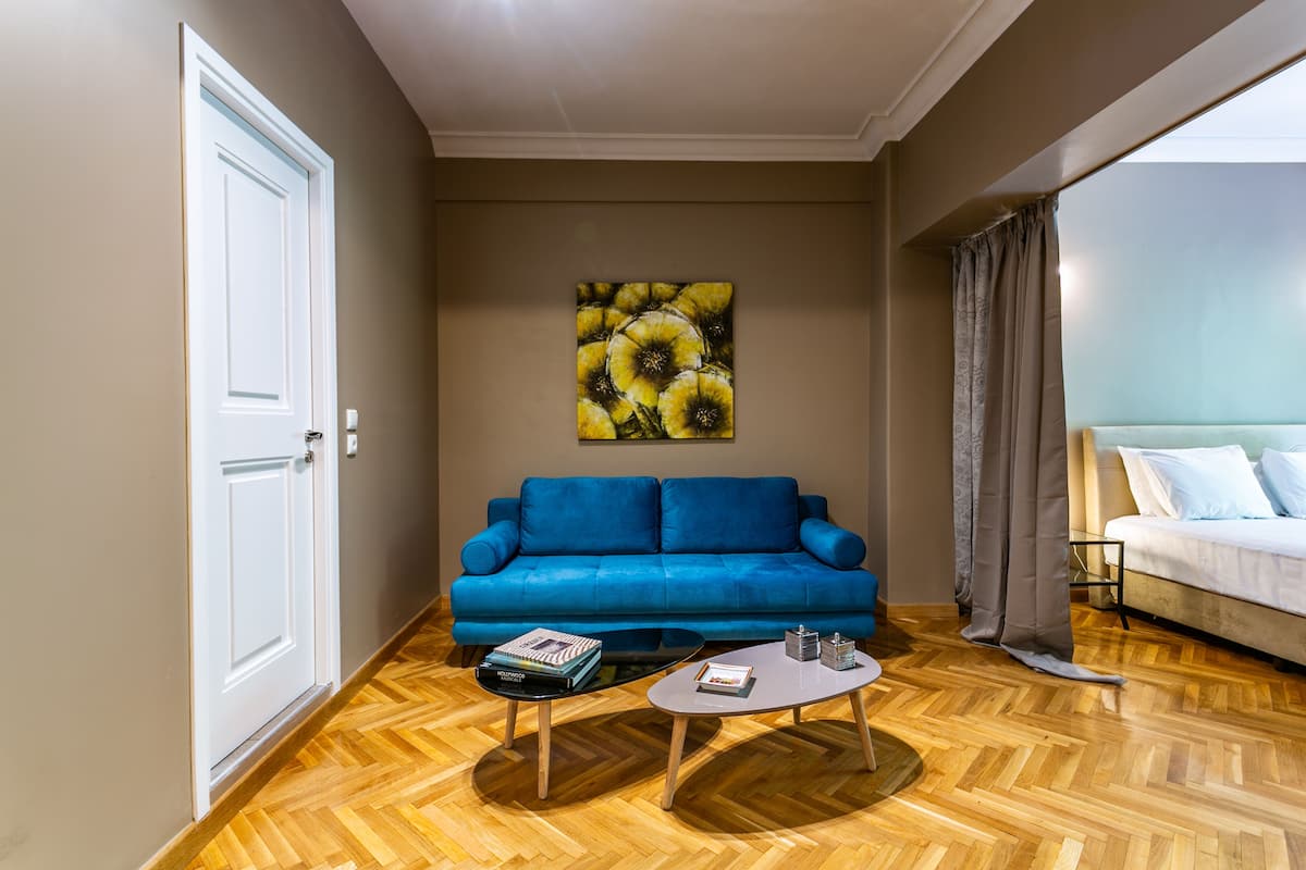 A room with a blue sofa, a coffee table, a painting on the wall, and a bed with a white headboard.