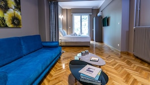 Een moderne hotelkamer met een blauwe bank, een houten visgraatvloer, een bed met witte lakens, een flatscreen televisie aan de muur en een groot raam met gordijnen.