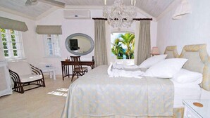 A spacious bedroom with a large bed, a sitting area with a chair and table, a mirror, and a view of palm trees through the window. - Emerald Beach 4 - Villa Plumbago (Gibbes)