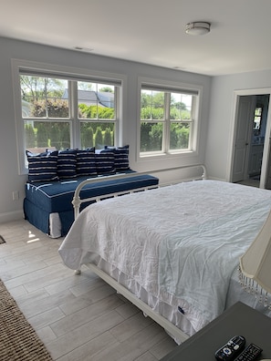 A bedroom with a large bed, a bench with pillows, and a view of greenery through the windows. - Maywood Bonnet Rails (Narragansett)