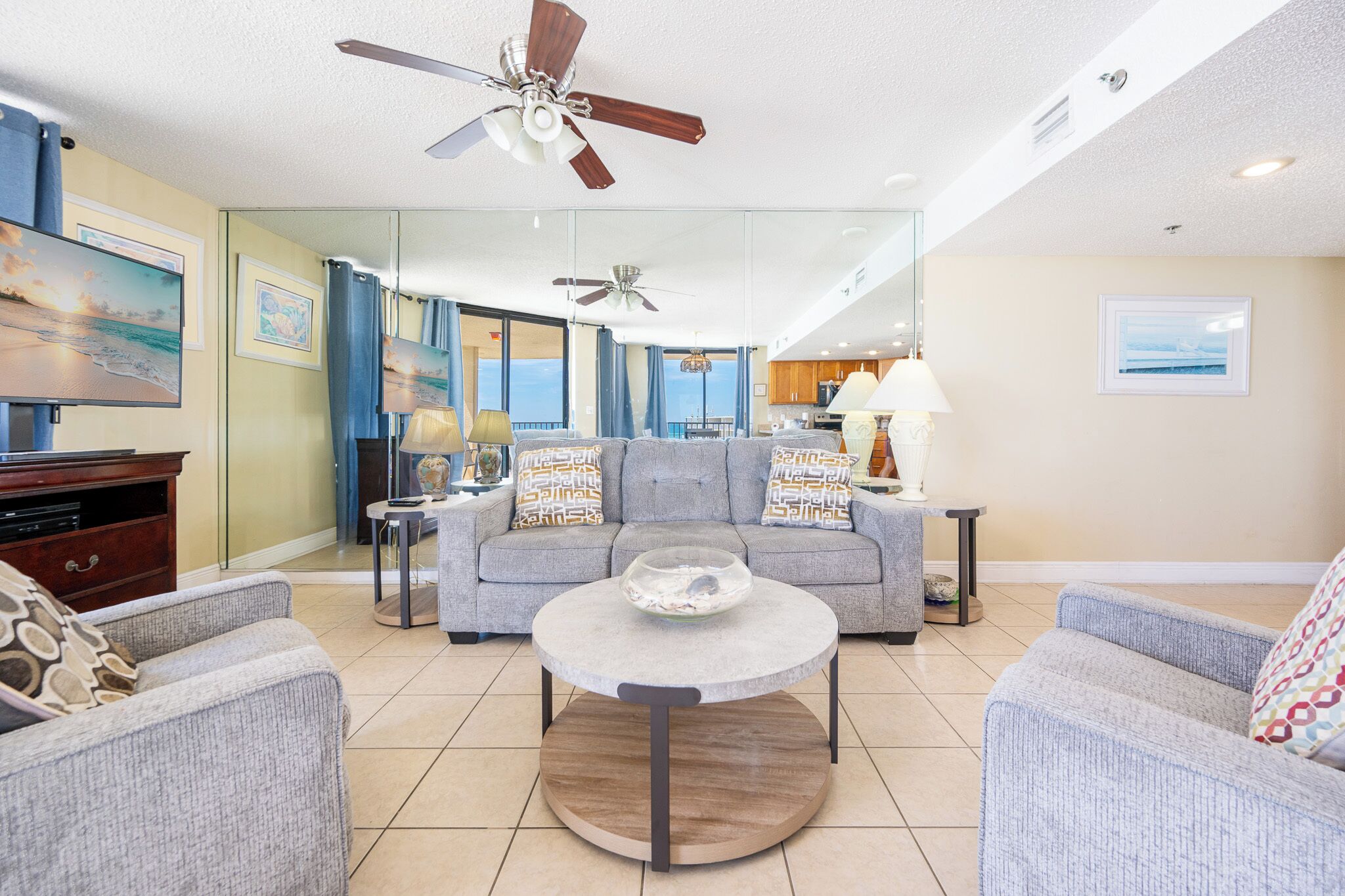 A living room with a sofa, armchairs, a coffee table, a TV, ceiling fans, and a kitchen in the background.