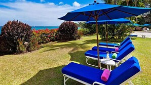 A poolside area with blue lounge chairs and umbrellas, a well-maintained lawn, and a view of the ocean.