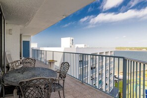 Un balcon avec une table ronde et des chaises, une balustrade, et une vue sur une étendue d’eau et des bâtiments au loin.