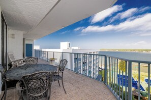Un balcon avec une table et des chaises, donnant sur une étendue d’eau et un terrain de tennis bleu.