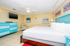 Un dormitorio con una cama, televisión, ventilador de techo, silla y obras de arte en las paredes.
