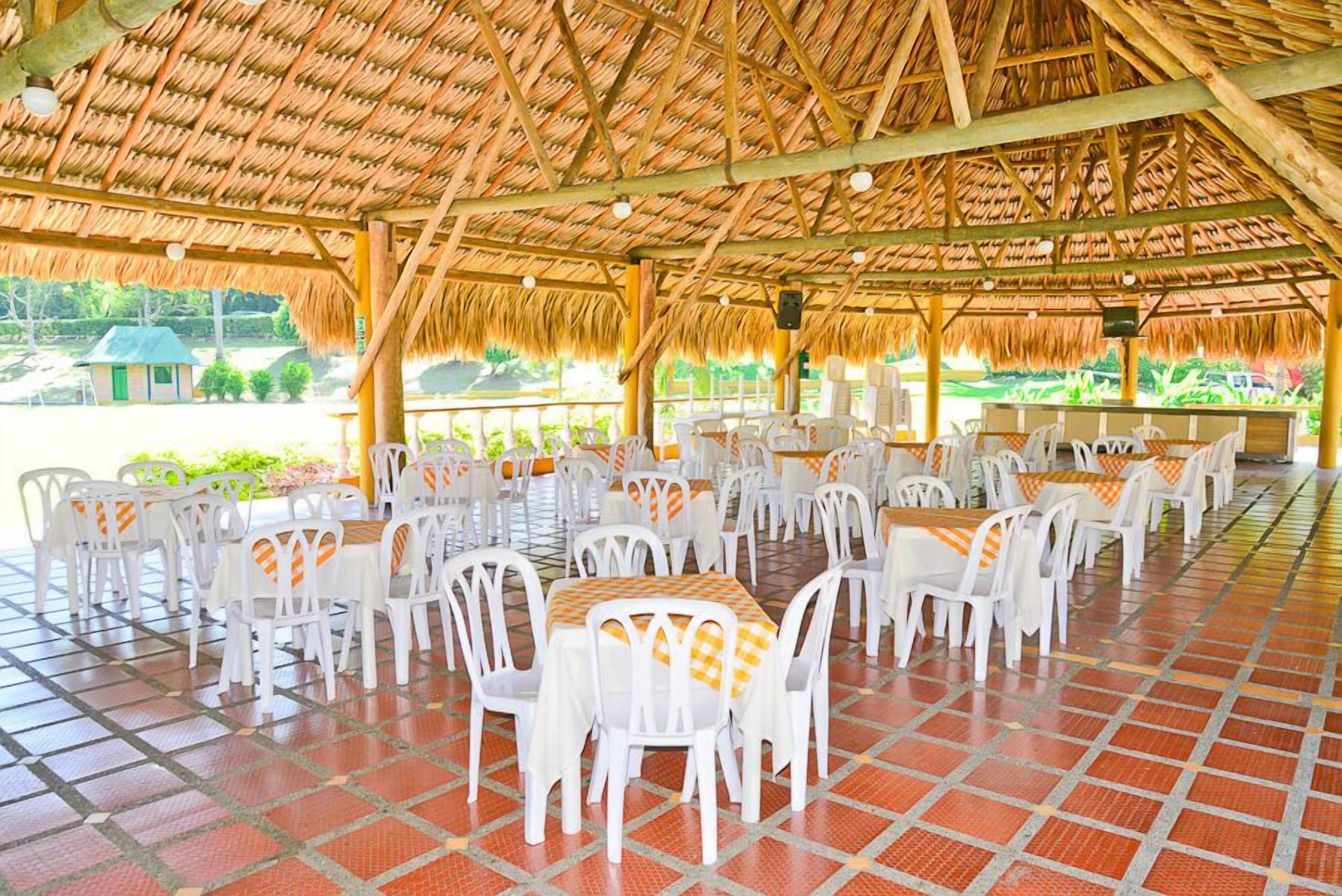 An outdoor dining area with white plastic chairs and tables, a thatched roof, and a tiled floor.
