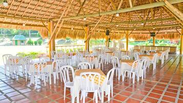 An outdoor dining area with white plastic chairs and tables, a thatched roof, and a tiled floor.