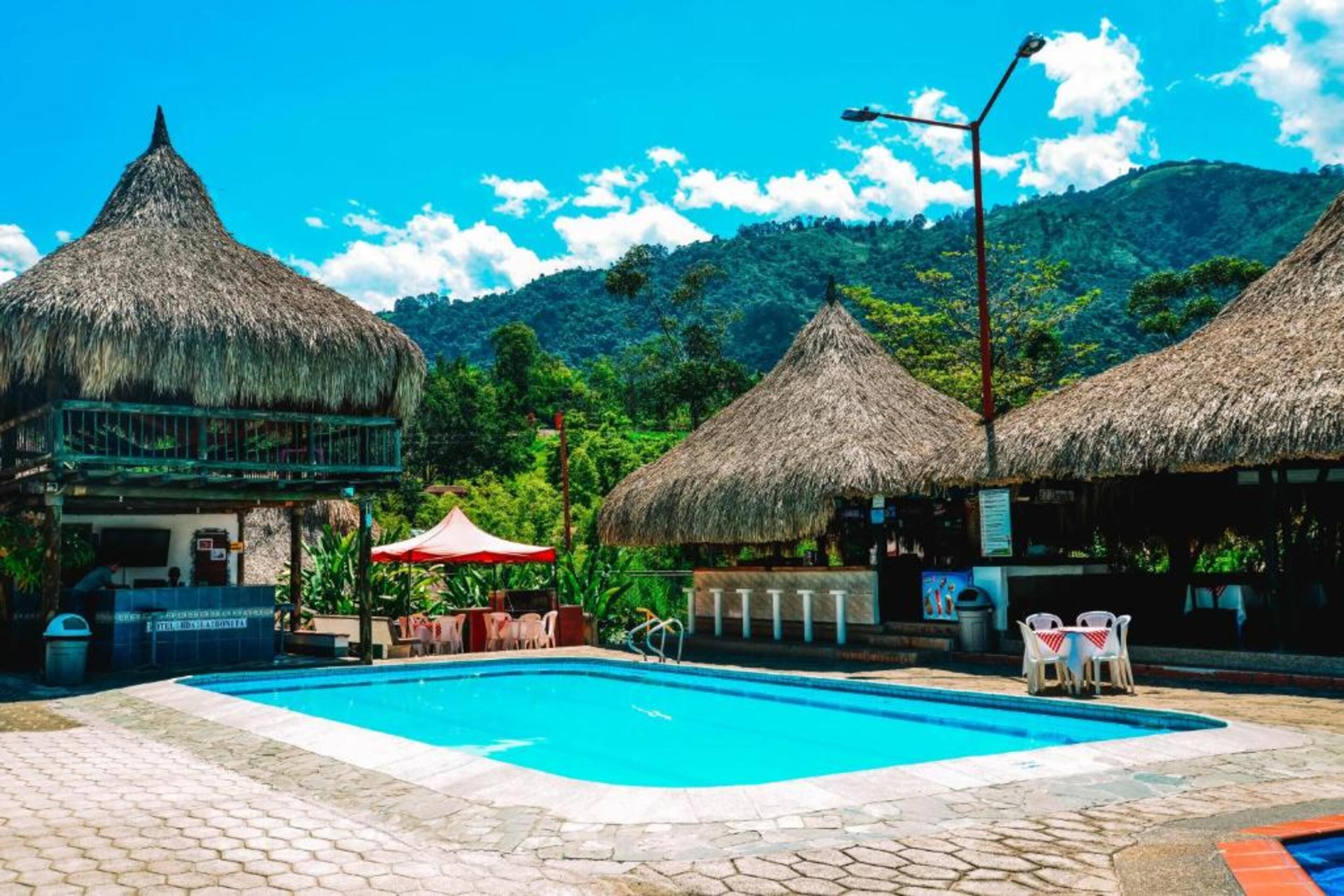 A swimming pool area with thatched-roof structures, a paved walkway, and a mountain backdrop.