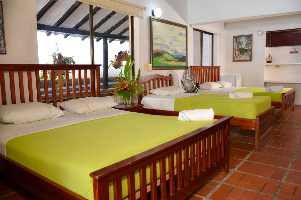 Una habitación de hotel con dos camas, cabecera de madera, colcha verde, una mesita con un florero y un cuadro en la pared.