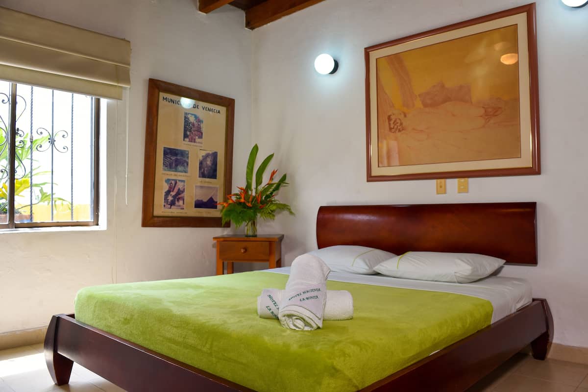 Una cama bien hecha con una colcha verde, un cuadro con una vista de la ciudad y una mesita con una planta.
