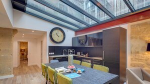 A modern kitchen with dark cabinets, a large clock, and a dining area with a table and chairs. - Sweett | Sentier II - Four Bedroom Apartment, Sleeps 9 (Paris)
