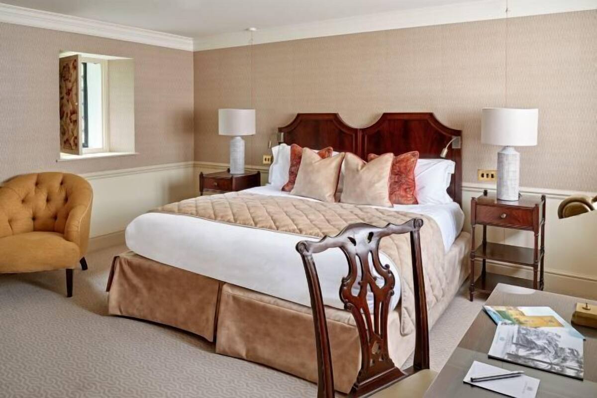 A hotel room with a large bed, a wooden headboard, two bedside tables with lamps, a chair, and a desk with a lamp and papers.