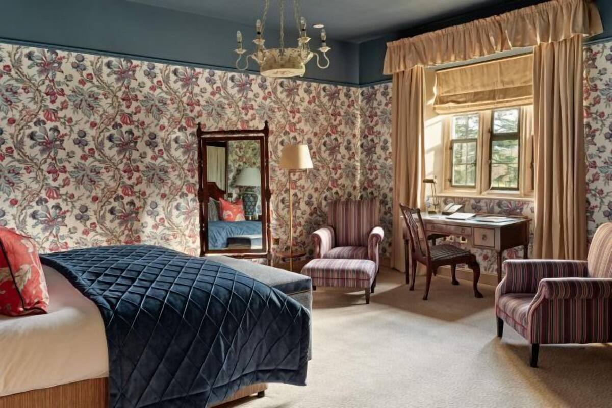 A bedroom with floral wallpaper, a bed with a quilted cover, a desk with a lamp, and two armchairs.