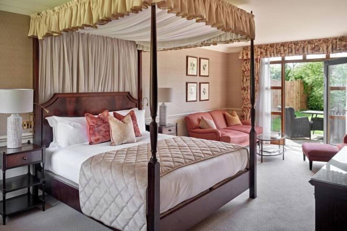 A four-poster canopy bed with a quilted duvet, bedside tables with lamps, a sofa, and a small round table.