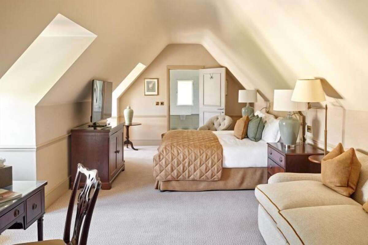 A bedroom with a sloped ceiling, a bed with a quilted cover, a bedside table with lamps, a dresser, and a chair.