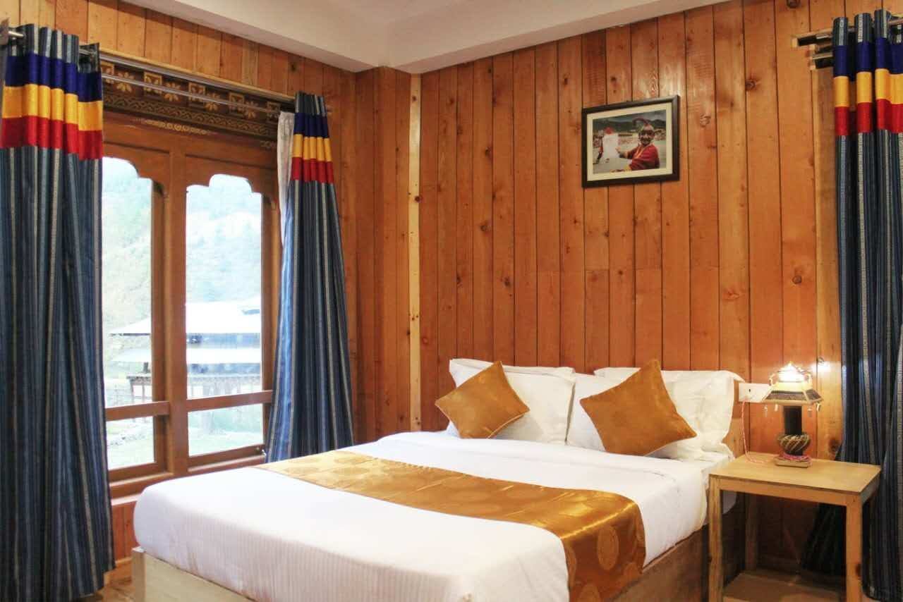 A wooden-paneled room with a bed, curtains, a framed picture on the wall, and a nightstand with a lamp.