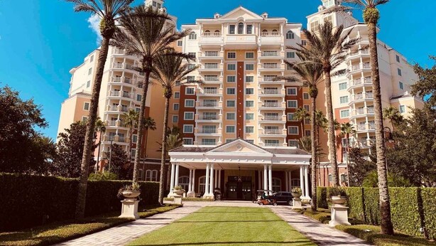 A large, multi-story hotel with a central entrance, surrounded by palm trees and a well-maintained lawn.