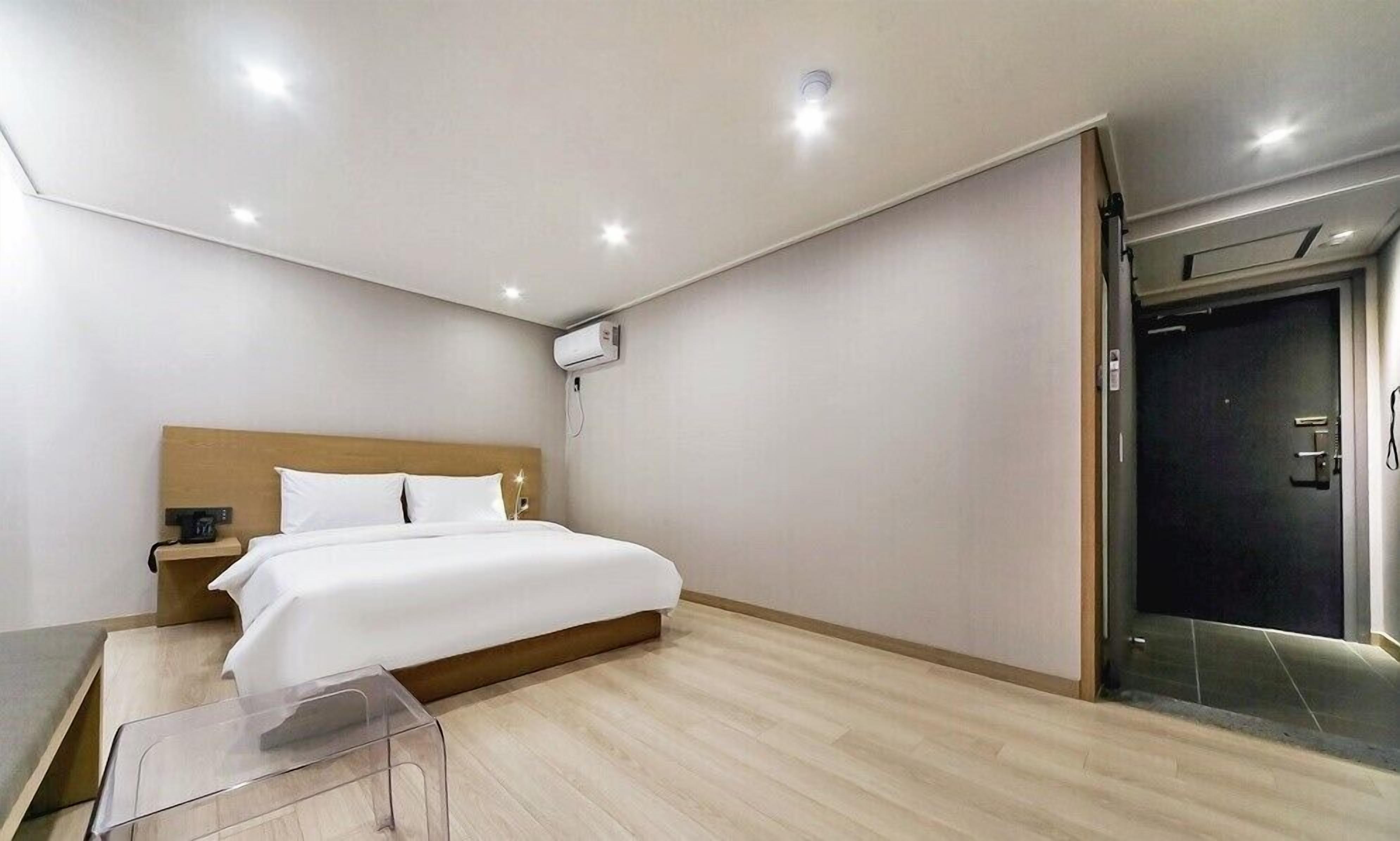 A modern hotel room with a wooden headboard bed, a bedside table with a lamp, a clear glass side table, and a dark door.