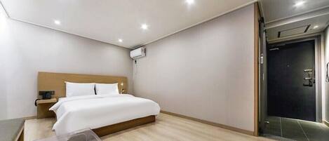 A modern hotel room with a wooden headboard bed, a bedside table with a lamp, a clear glass side table, and a dark door.
