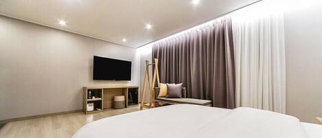 A modern hotel room with a large bed, a flat-screen TV mounted on the wall, a wooden side table, and a large window with curtains.