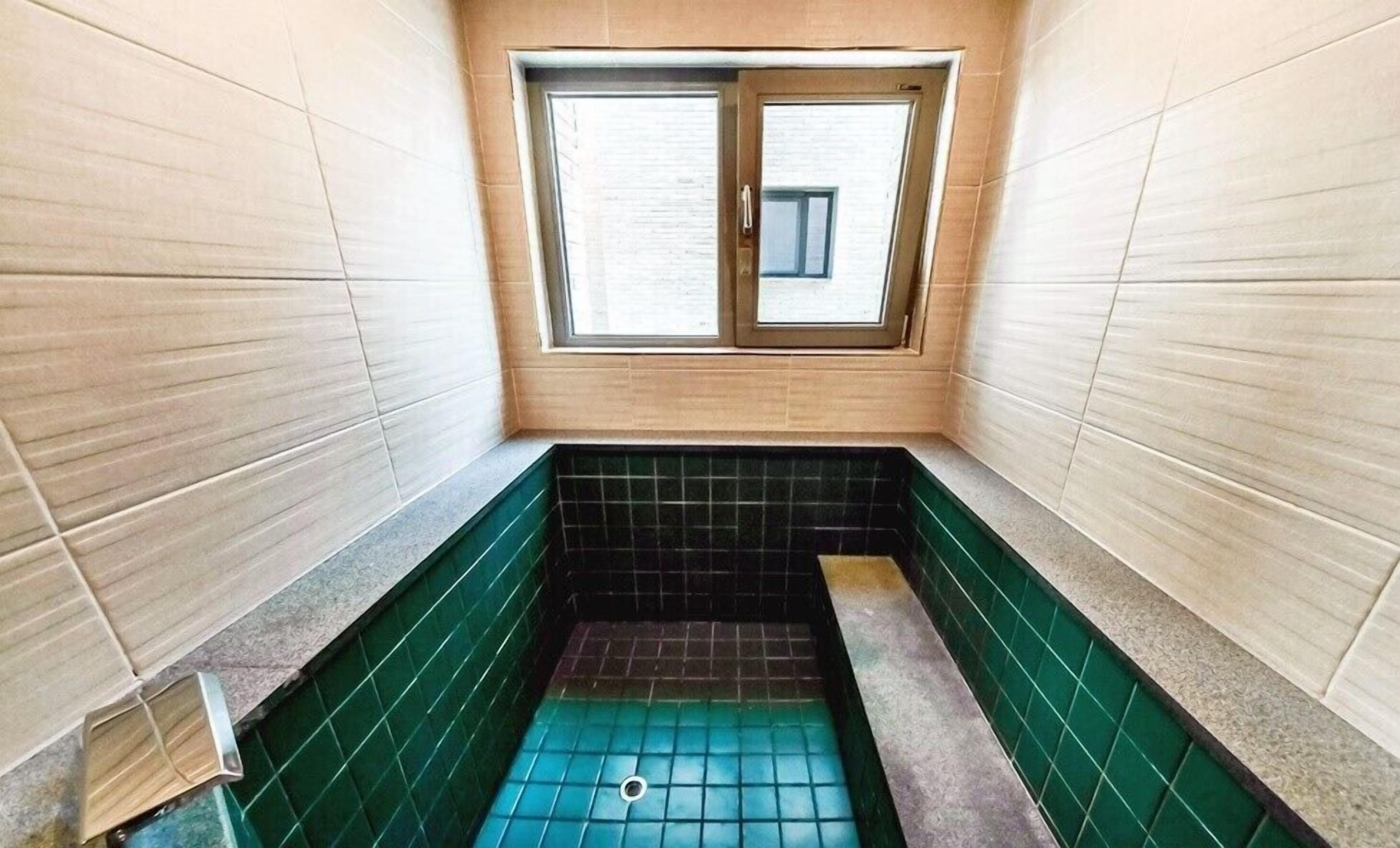 Un baño pequeño y estrecho con ventana, piso de baldosas verde y negro, y paredes de color claro.