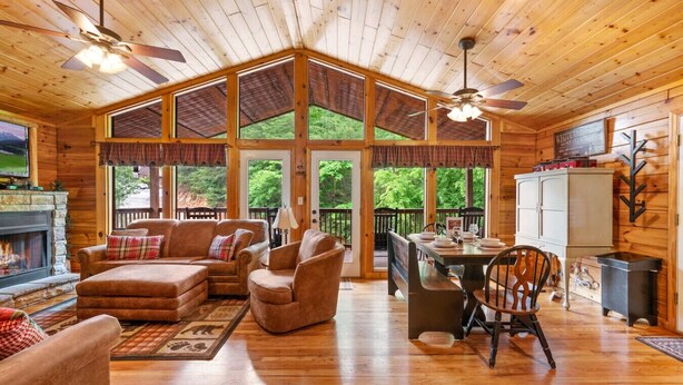 A cozy cabin interior with a fireplace, wooden furniture, and large windows offering a view of the outdoors. - Best Location/HotTub/Resort Pool/Game Rm/Fast WiFi
Dollywood 2 minutes away! (Sevierville)