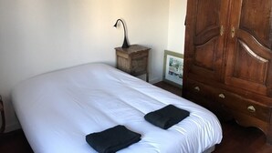 A neatly made bed with white sheets and two black pillows, a small wooden nightstand with a lamp, and a wooden wardrobe with brass handles. - Pleasant house on the Arcachon basin (La Teste-de-Buch)