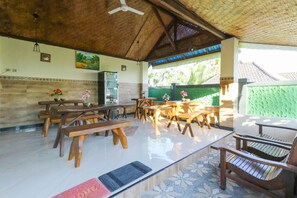 A covered outdoor dining area with wooden tables and benches, a ceiling fan, and a view of greenery outside. - Sri Gunadi Bungalow (Penida Island)