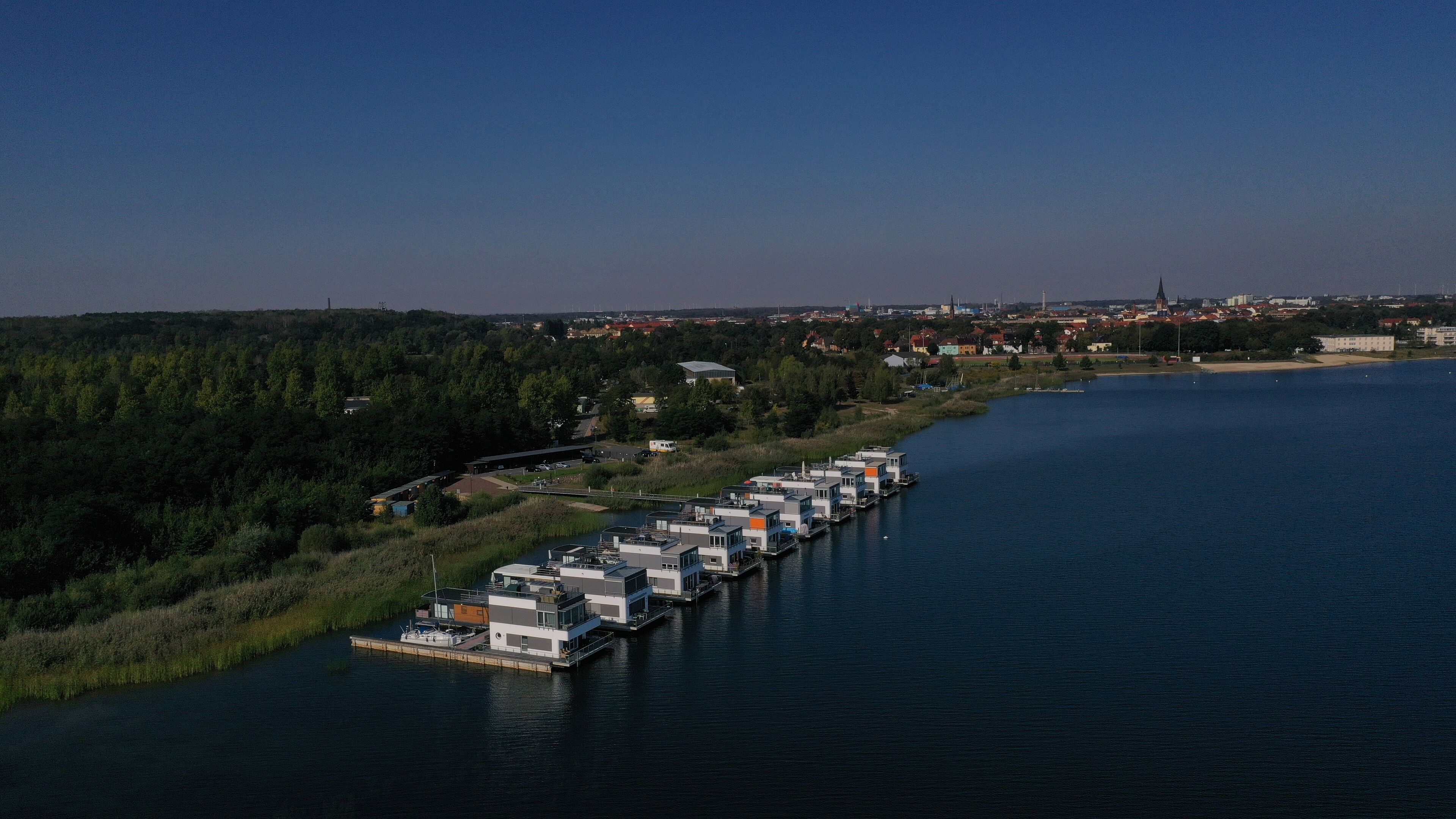 Una hilera de edificios modernos de varios pisos, con un muelle que se adentra en un lago tranquilo, rodeado de un bosque denso y un pueblo distante.