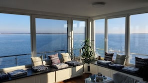 A room with large windows overlooking a body of water, featuring a seating area with cushions and a plant.