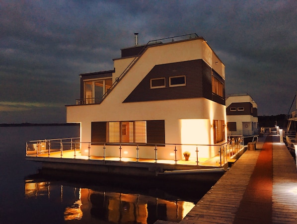 A modern houseboat with a flat roof, large windows, and a dock extending into calm water.