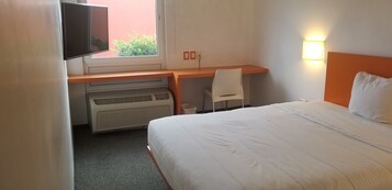 Habitación de hotel con cama, escritorio con silla, televisor y ventana con vista a follaje.