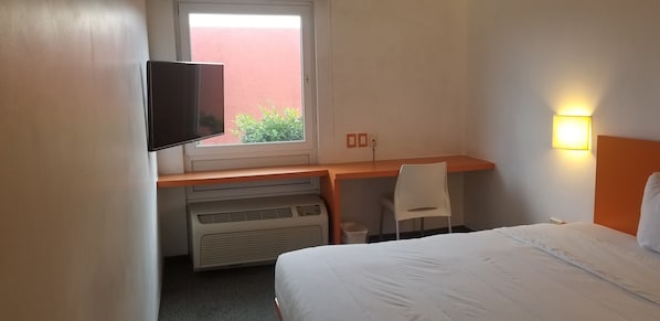 A hotel room with a bed, a desk with a chair, a television, a window, and a wall-mounted light.