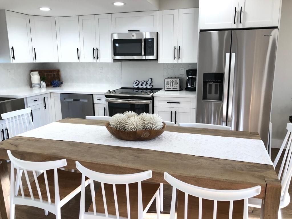Une cuisine moderne avec des meubles blancs, des appareils en acier inoxydable et une table à manger en bois, assortie de chaises blanches.
