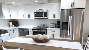 Une cuisine moderne avec des meubles blancs, des appareils en acier inoxydable et une table à manger en bois, assortie de chaises blanches.