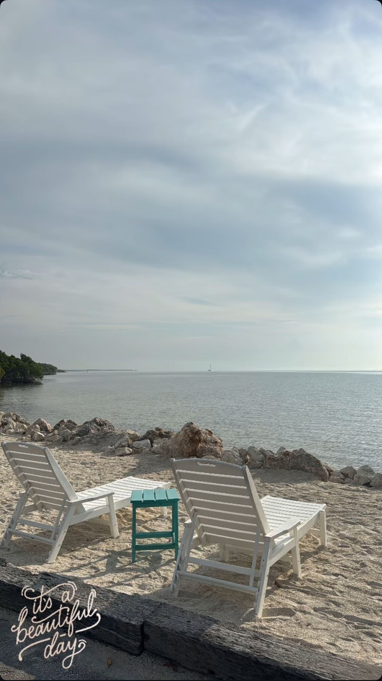 Une plage avec des chaises longues blanches et une table verte, un rivage rocheux et une mer calme sous un ciel nuageux.
